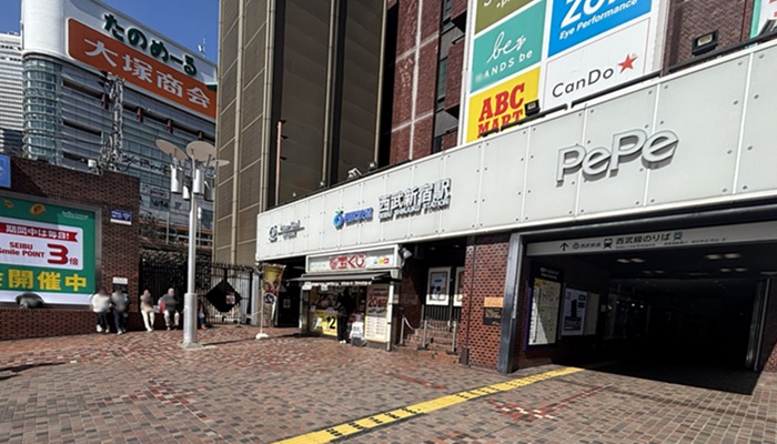 西武新宿駅の正面入口とPePeのロゴ。左側のレンガ壁沿いに座って待つ人たちの様子。