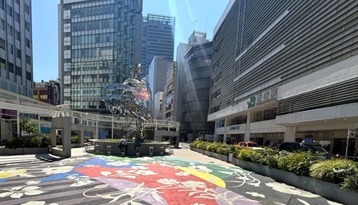 新宿駅東口の「花尾さん広場」。地面に描かれた花のアートと、銀色の巨大オブジェ「花尾」、右側に見えるJR新宿駅の駅舎。