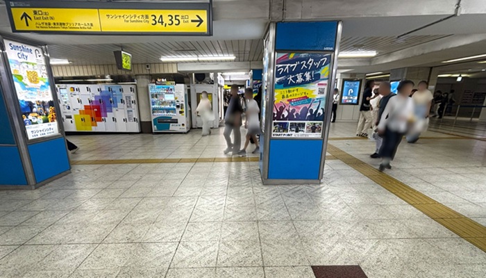 池袋駅地下通路。天井に『サンシャインシティ方面』、『出口 34, 35 →』と書かれた黄色い案内看板があり、青い広告柱が並ぶ通路